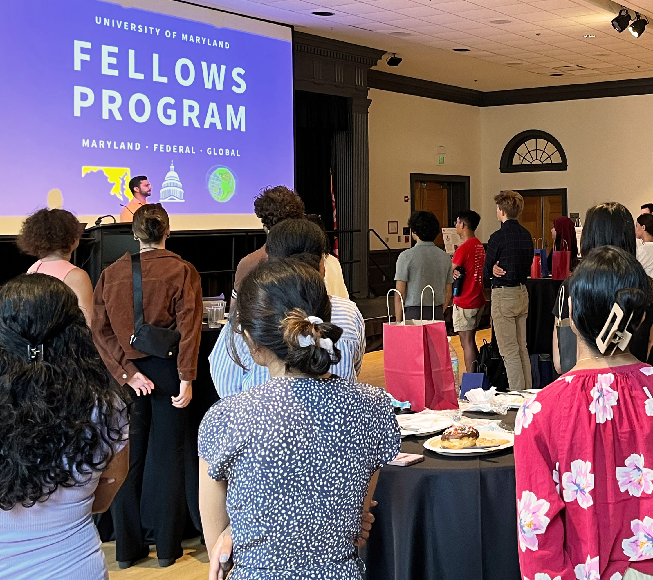 Many diverse new Fellows standing in a ballroom and listening to Jonathan Lee speak on a stage in fron of the UMD Fellows Program Logo